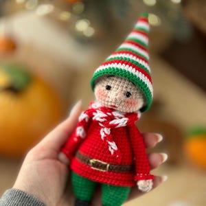 May include: A crocheted elf doll wearing a red and white striped hat, a red sweater, a white and red scarf, and green pants.