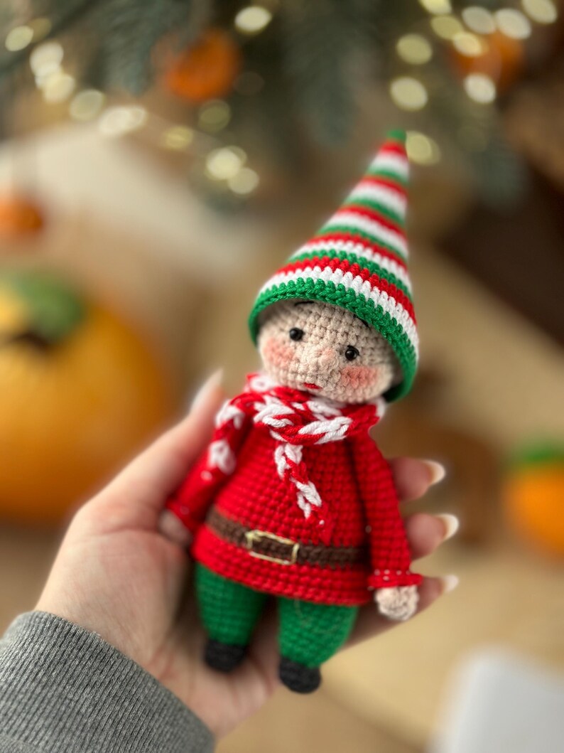 May include: A crocheted elf doll wearing a red and white striped hat, a red sweater, a white and red scarf, and green pants.