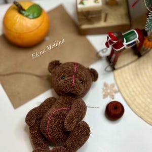 以下が含まれることがあります： 茶色の毛糸で編まれたテディベアで、ピンク色の糸が体全体に縫い付けられています。ベアは白い表面に置かれており、茶色の毛糸で編まれた雪の結晶と茶色の糸巻きがあります。