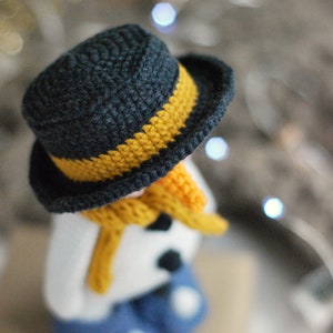 May include: A crocheted snowman wearing a black and yellow hat, a yellow scarf, and blue pants. The snowman is sitting on a brown surface.