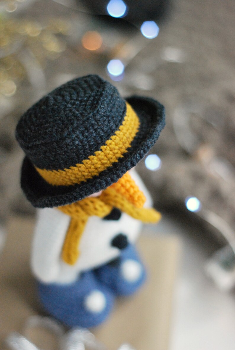 May include: A crocheted snowman wearing a black and yellow hat, a yellow scarf, and blue pants. The snowman is sitting on a brown surface.