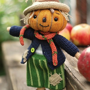 May include: A crocheted pumpkin scarecrow doll wearing a blue sweater, green striped trousers, and a brown hat. The doll is holding a red apple.