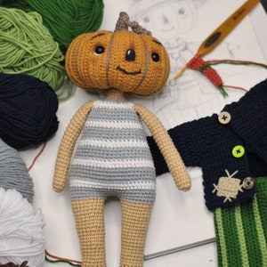 May include: A crocheted pumpkin-headed doll wearing a striped shirt and brown shoes. The doll is surrounded by balls of yarn in various colours, including green, blue, and brown. There is also a crocheted blue cardigan with green and white striped trousers and a crocheted hat.