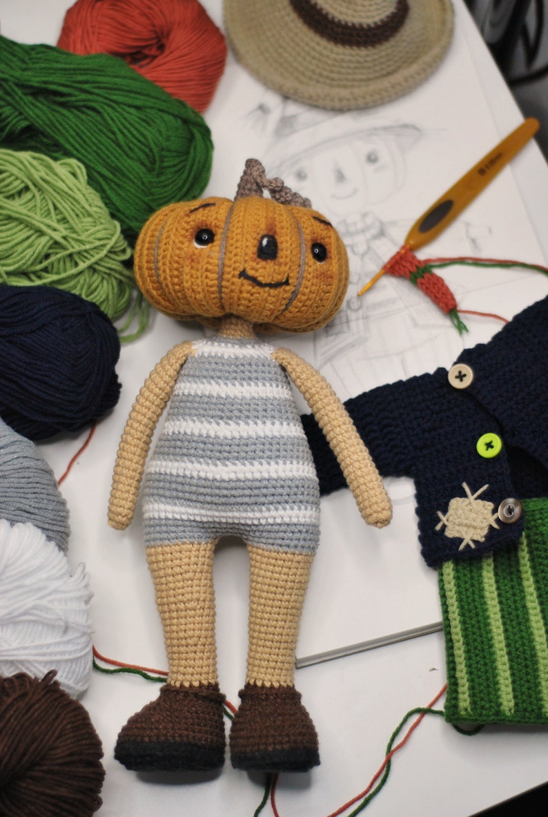 May include: A crocheted pumpkin-headed doll wearing a striped shirt and brown shoes. The doll is surrounded by balls of yarn in various colours, including green, blue, and brown. There is also a crocheted blue cardigan with green and white striped trousers and a crocheted hat.