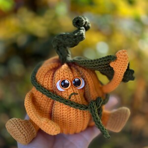 May include: A crocheted pumpkin with a sad face and a green stem. The pumpkin is wearing a green scarf around its neck.