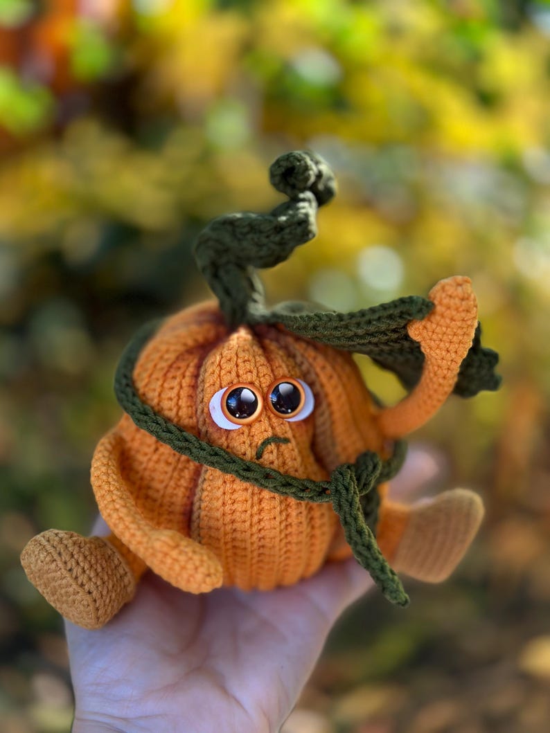 May include: A crocheted pumpkin with a sad face and a green stem. The pumpkin is wearing a green scarf around its neck.
