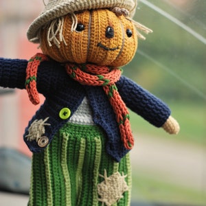 May include: A crocheted pumpkin-headed scarecrow doll wearing a blue jacket, green trousers, and a red and green scarf. The doll has a friendly face and is holding out its arms.