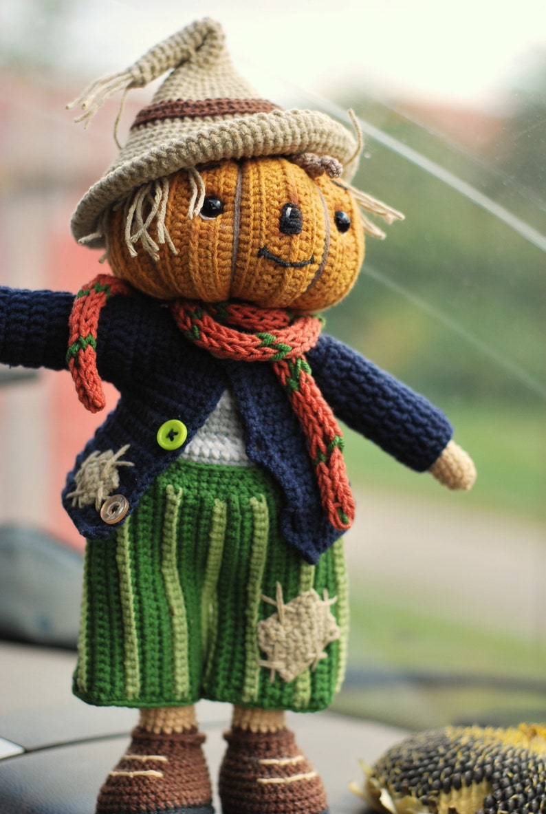 May include: A crocheted pumpkin-headed scarecrow doll wearing a blue jacket, green trousers, and a red and green scarf. The doll has a friendly face and is holding out its arms.