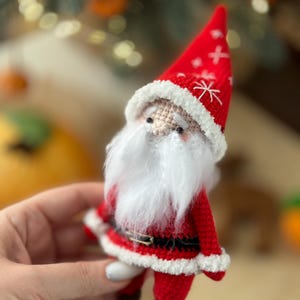May include: A crocheted Santa Claus ornament with a red hat, white beard, and a black belt. The ornament is holding a small white bag.