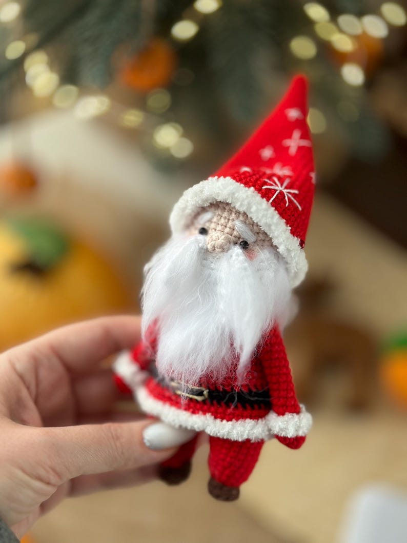 May include: A crocheted Santa Claus ornament with a red hat, white beard, and a black belt. The ornament is holding a small white bag.