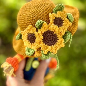 May include: A golden yellow crochet hat decorated with three sunflowers. The sunflowers have yellow petals, brown centres, and green leaves. The hat has a brim and a rounded crown, suitable for a child's costume or decorative item.