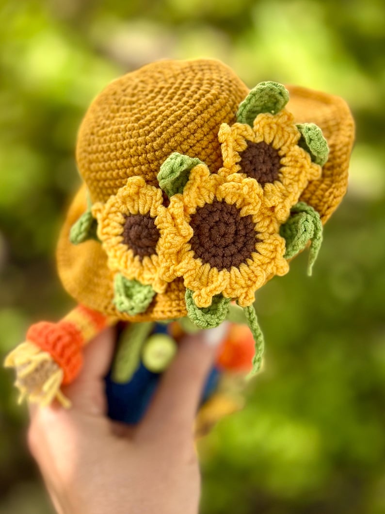 May include: A golden yellow crochet hat decorated with three sunflowers. The sunflowers have yellow petals, brown centres, and green leaves. The hat has a brim and a rounded crown, suitable for a child's costume or decorative item.
