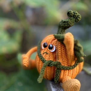May include: A crocheted pumpkin toy with a green stem and a green belt. The pumpkin has a sad expression with two white eyes and a small mouth.