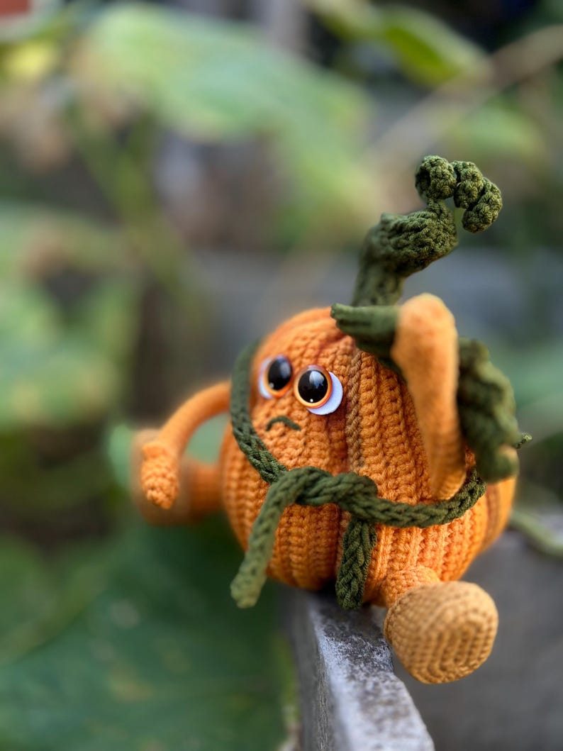 May include: A crocheted pumpkin toy with a green stem and a green belt. The pumpkin has a sad expression with two white eyes and a small mouth.