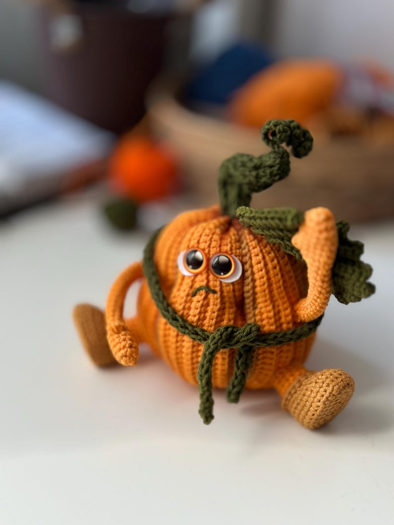 May include: A crocheted pumpkin with a green stem and a green crocheted belt. The pumpkin has a sad expression with two large eyes.