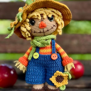 May include: A handmade crochet scarecrow doll wearing a straw hat, green scarf, and blue dungarees. The doll has a stitched smile, orange nose, and a sunflower accent. The doll is approximately 20 cm tall.
