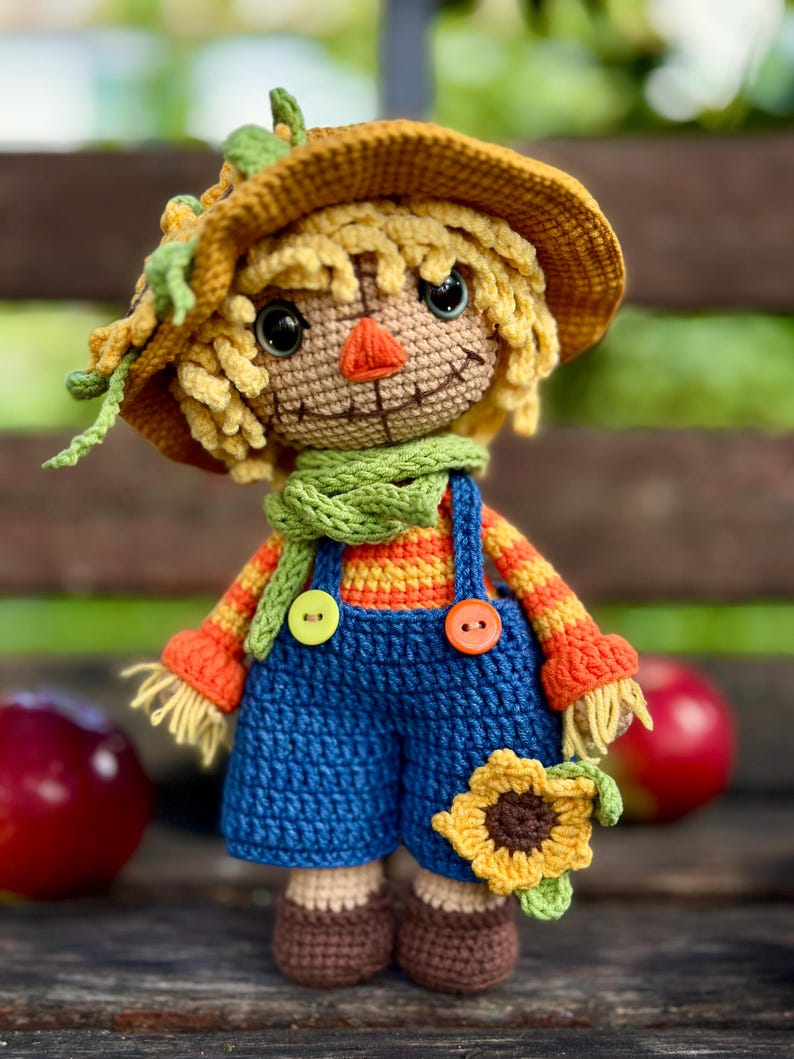 May include: A handmade crochet scarecrow doll wearing a straw hat, green scarf, and blue dungarees. The doll has a stitched smile, orange nose, and a sunflower accent. The doll is approximately 20 cm tall.