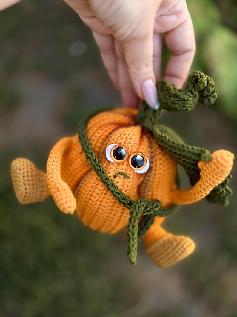 May include: A crocheted pumpkin toy with a green stem and large eyes. The pumpkin is orange and has a sad expression. The toy is being held by a hand.