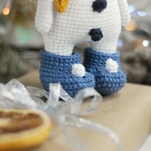 May include: A white crocheted snowman with black buttons and a yellow carrot nose. The snowman is wearing blue crocheted boots with white pom-poms.