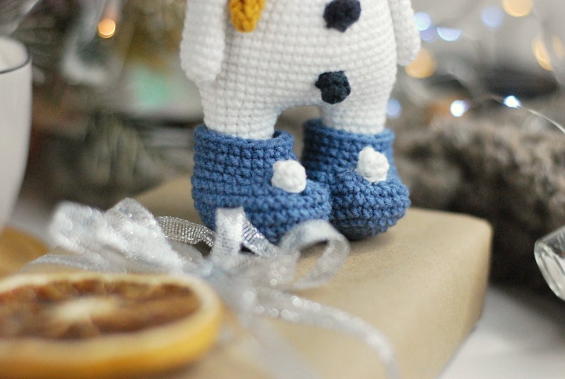 May include: A white crocheted snowman with black buttons and a yellow carrot nose. The snowman is wearing blue crocheted boots with white pom-poms.