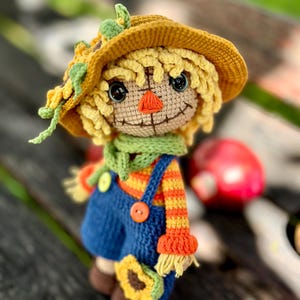 May include: A handmade crochet scarecrow doll. It features a yellow hat with green leaves and a sunflower. The doll has a smiling face, a green scarf, and blue dungarees with orange and yellow striped sleeves. A sunflower is attached to the dungarees.