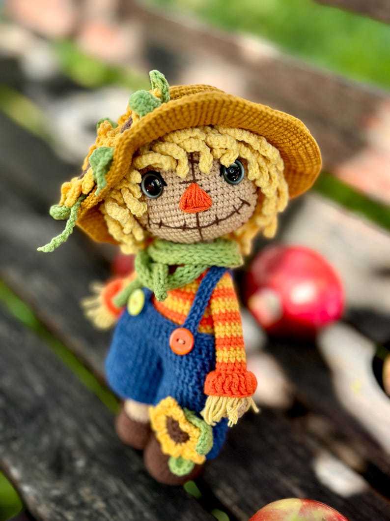 May include: A handmade crochet scarecrow doll. It features a yellow hat with green leaves and a sunflower. The doll has a smiling face, a green scarf, and blue dungarees with orange and yellow striped sleeves. A sunflower is attached to the dungarees.