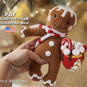 Puede incluir: Patrón de crochet PDF para un juguete amigurumi en forma de hombre de jengibre. El hombre de jengibre es marrón con detalles de glaseado blanco y sostiene una taza roja con malvaviscos blancos y un bastón de caramelo.