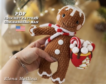 Puede incluir: Patrón de crochet PDF para un juguete amigurumi en forma de hombre de jengibre. El hombre de jengibre es marrón con detalles de glaseado blanco y sostiene una taza roja con malvaviscos blancos y un bastón de caramelo.