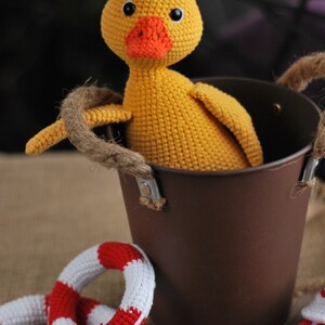 May include: A yellow crocheted duck toy sitting in a brown metal bucket with rope handles. There are two red and white crocheted life preserver rings on the table.