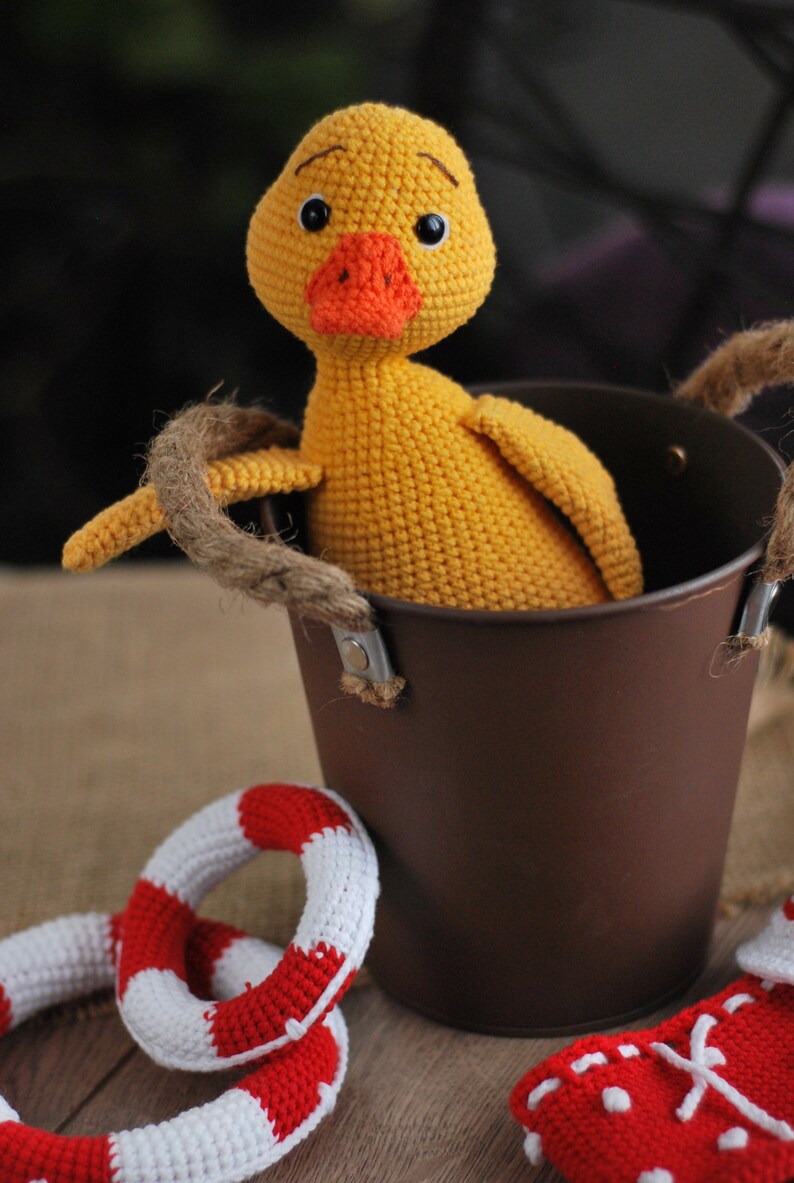 May include: A yellow crocheted duck toy sitting in a brown metal bucket with rope handles. There are two red and white crocheted life preserver rings on the table.
