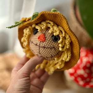 May include: A handmade crochet scarecrow head with a yellow hat and curly hair. The scarecrow has black eyes, an orange nose, and a stitched smile. The hat has green leaf accents.