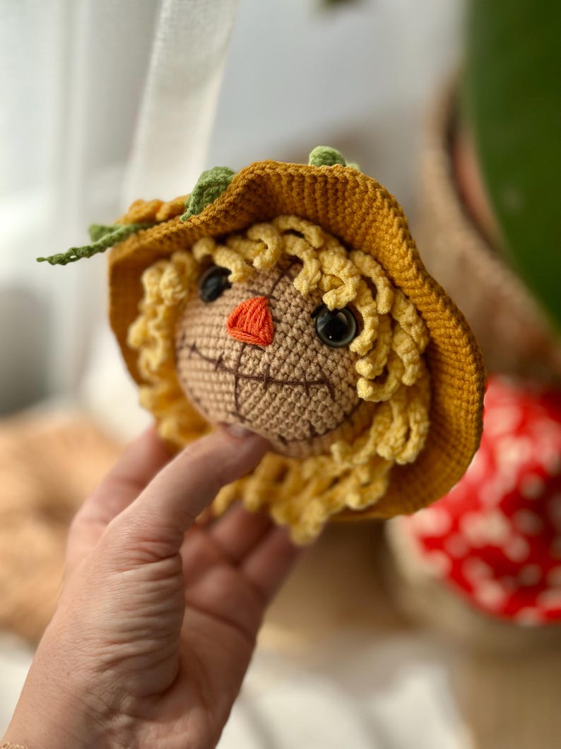 May include: A handmade crochet scarecrow head with a yellow hat and curly hair. The scarecrow has black eyes, an orange nose, and a stitched smile. The hat has green leaf accents.