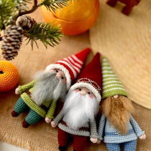 May include: Three crocheted gnomes with different colored hats and beards. The gnome on the left is wearing a green and brown striped hat with a white beard. The gnome in the middle is wearing a red and white striped hat with a white beard. The gnome on the right is wearing a green and white striped hat with a brown beard.