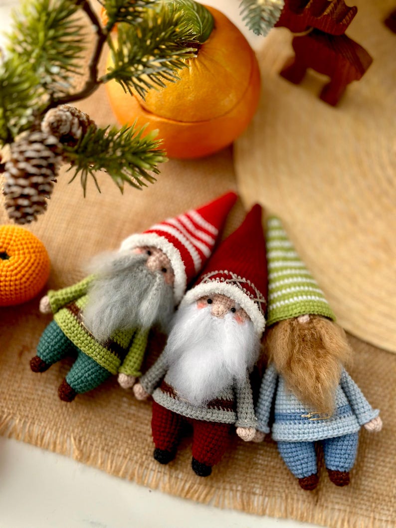 May include: Three crocheted gnomes with different colored hats and beards. The gnome on the left is wearing a green and brown striped hat with a white beard. The gnome in the middle is wearing a red and white striped hat with a white beard. The gnome on the right is wearing a green and white striped hat with a brown beard.