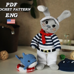 Könnte beinhalten: Häkelmuster-PDF für einen grauen Hasen, der ein Matrosen-Outfit trägt, ein blau-weiß gestreiftes Hemd, einen roten Schal und einen weißen Hut. Der Hase hält einen blauen gehäkelten Delfin mit einem roten und weißen Rettungsring. Es gibt auch eine blaue gehäkelte Tasche mit einem weißen Riemen.