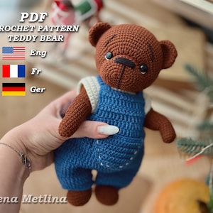 Puede incluir: Patrón de crochet PDF para un osito de peluche con un mono azul. El patrón está disponible en inglés, francés y alemán. El oso es marrón con ojos negros y una nariz negra.