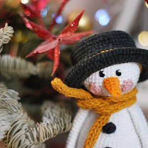 May include: A crocheted snowman ornament with a black hat, yellow scarf, and white face. The snowman is surrounded by a green and white pine branch.