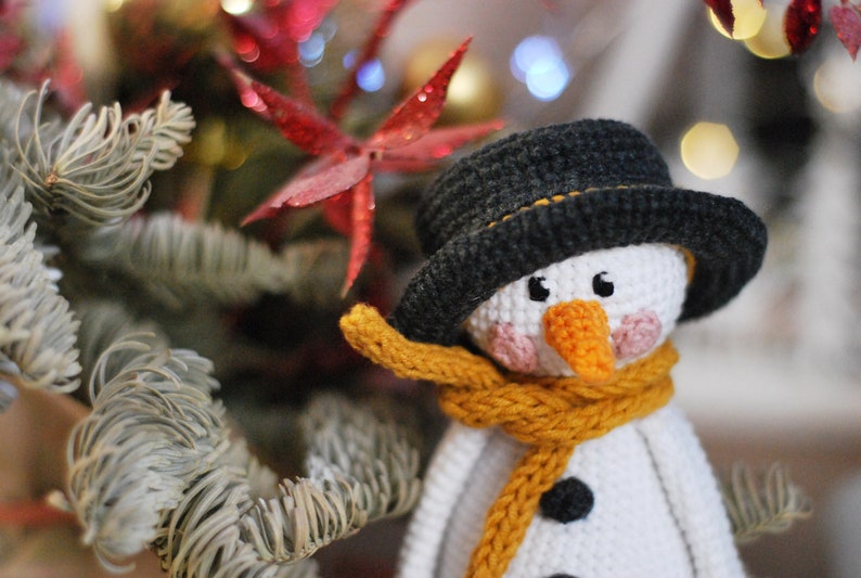 May include: A crocheted snowman ornament with a black hat, yellow scarf, and white face. The snowman is surrounded by a green and white pine branch.