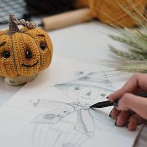 May include: A person is sketching a scarecrow with a pencil on a white sheet of paper. A crocheted pumpkin with a smiling face is sitting on the table next to the paper.