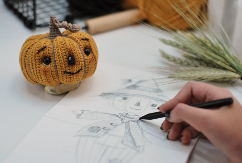May include: A person is sketching a scarecrow with a pencil on a white sheet of paper. A crocheted pumpkin with a smiling face is sitting on the table next to the paper.