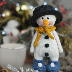 May include: A crocheted snowman toy with a black hat, yellow scarf, and blue boots. The snowman has a white body, black eyes, a black button nose, and black buttons for eyes.