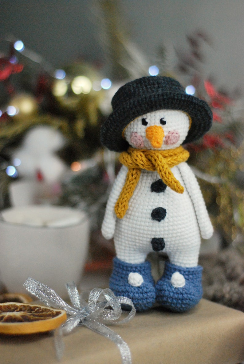 May include: A crocheted snowman toy with a black hat, yellow scarf, and blue boots. The snowman has a white body, black eyes, a black button nose, and black buttons for eyes.