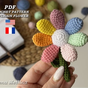 May include: A colorful crocheted flower keychain with a green stem. The flower has seven petals in different colors: pink, light pink, blue, light green, orange, yellow, and white. The text "PDF CROCHET PATTERN KEYCHAIN FLOWER" is visible in the image.