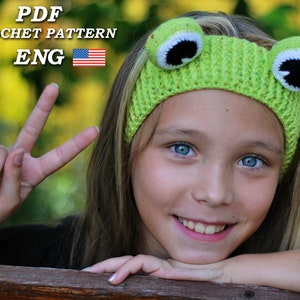 Puede incluir: Una niña pequeña con un diadema verde de ganchillo con ojos de rana. La diadema es verde con detalles blancos y negros. La niña sonríe y tiene el pelo rubio.