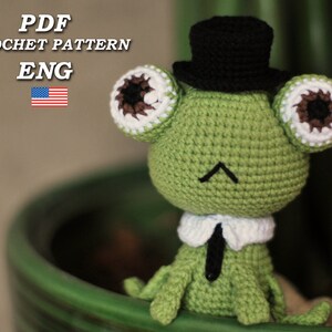 May include: Crochet pattern for a green frog wearing a black top hat and a white collar. The pattern is available in English.