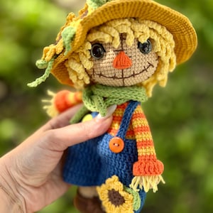 May include: A handmade crochet scarecrow doll with a yellow hat, straw-like hair, and a green scarf. The doll wears blue dungarees with an orange button and a striped orange and yellow top. A sunflower is attached to the doll's dungarees.