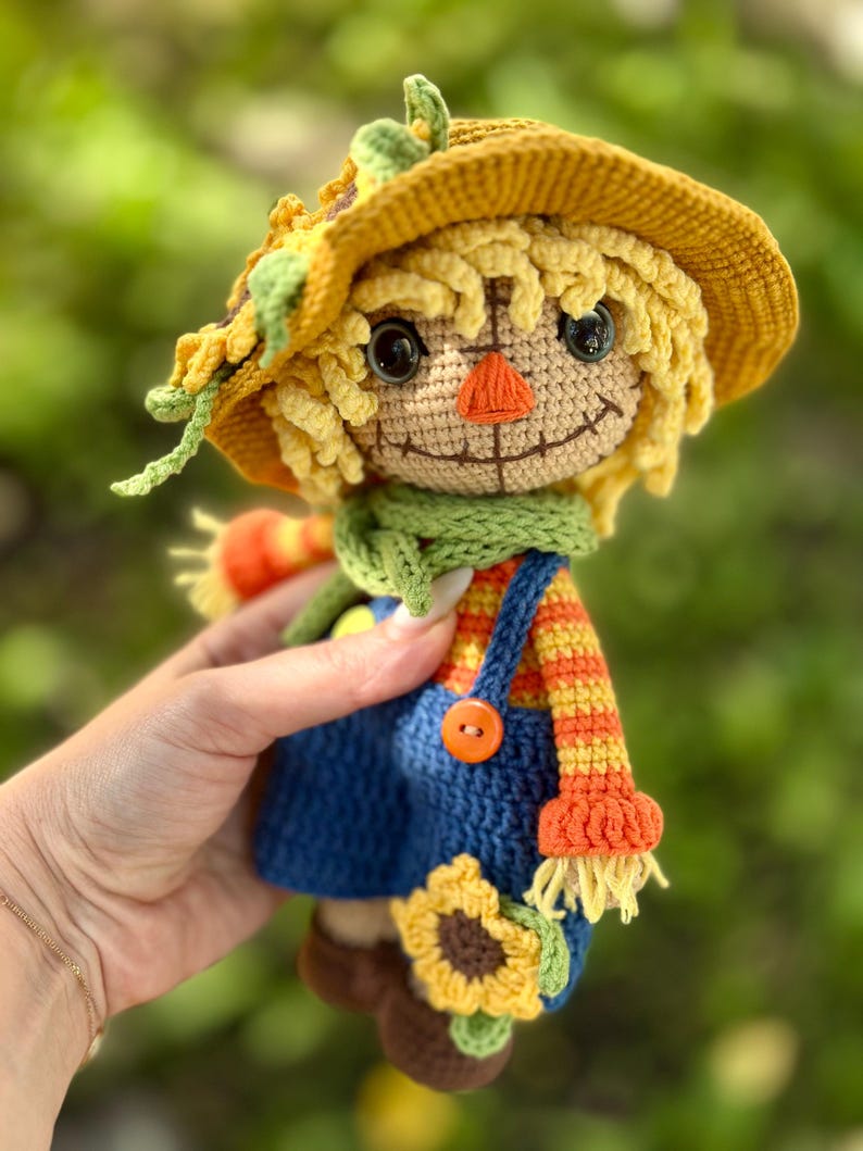 May include: A handmade crochet scarecrow doll with a yellow hat, straw-like hair, and a green scarf. The doll wears blue dungarees with an orange button and a striped orange and yellow top. A sunflower is attached to the doll's dungarees.