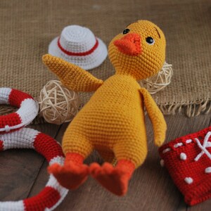 May include: A yellow crocheted duck toy wearing red and white crocheted swim trunks. The duck is lying on a wooden surface with a white crocheted hat, a woven basket, and two red and white crocheted life preservers.