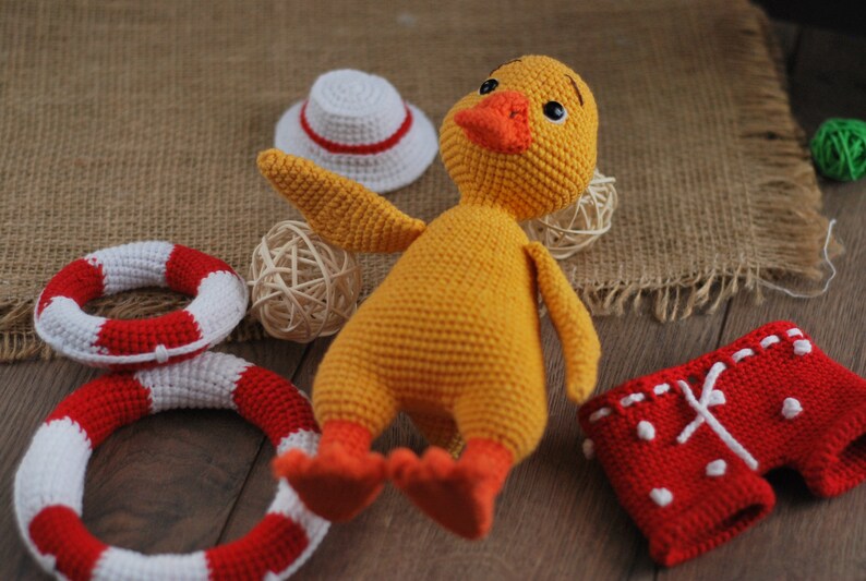 May include: A yellow crocheted duck toy wearing red and white crocheted swim trunks. The duck is lying on a wooden surface with a white crocheted hat, a woven basket, and two red and white crocheted life preservers.