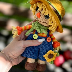 May include: A handmade crochet scarecrow doll with a yellow hat, straw hair, and a green scarf. The doll wears blue dungarees with orange and yellow striped sleeves. It holds a small sunflower. The doll is approximately 15 cm tall.